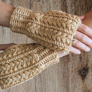 May include: A pair of tan crochet fingerless gloves with a cable knit design. The gloves are made of a soft yarn and are designed to keep hands warm while leaving fingers free. The gloves are shown on a wooden surface.