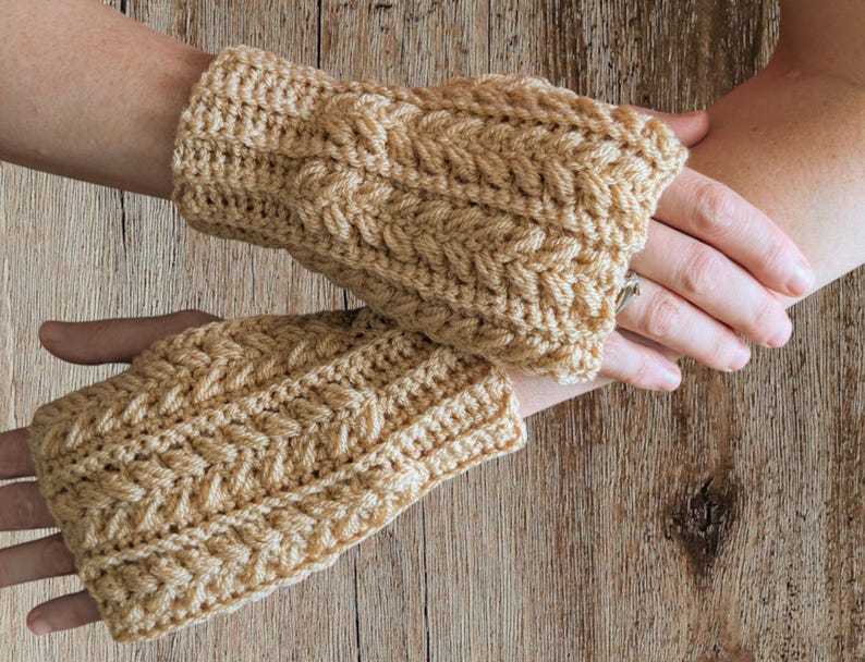 May include: A pair of tan crochet fingerless gloves with a cable knit design. The gloves are made of a soft yarn and are designed to keep hands warm while leaving fingers free. The gloves are shown on a wooden surface.
