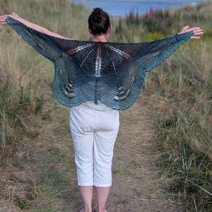 Puede incluir: Una persona lleva un chal hecho a mano, de tejido abierto, con un dise&ntilde;o de mariposa. El chal hace una transici&oacute;n de negro a verde azulado, con un patr&oacute;n delicado y calado. El fondo presenta un paisaje costero con dunas y el mar.