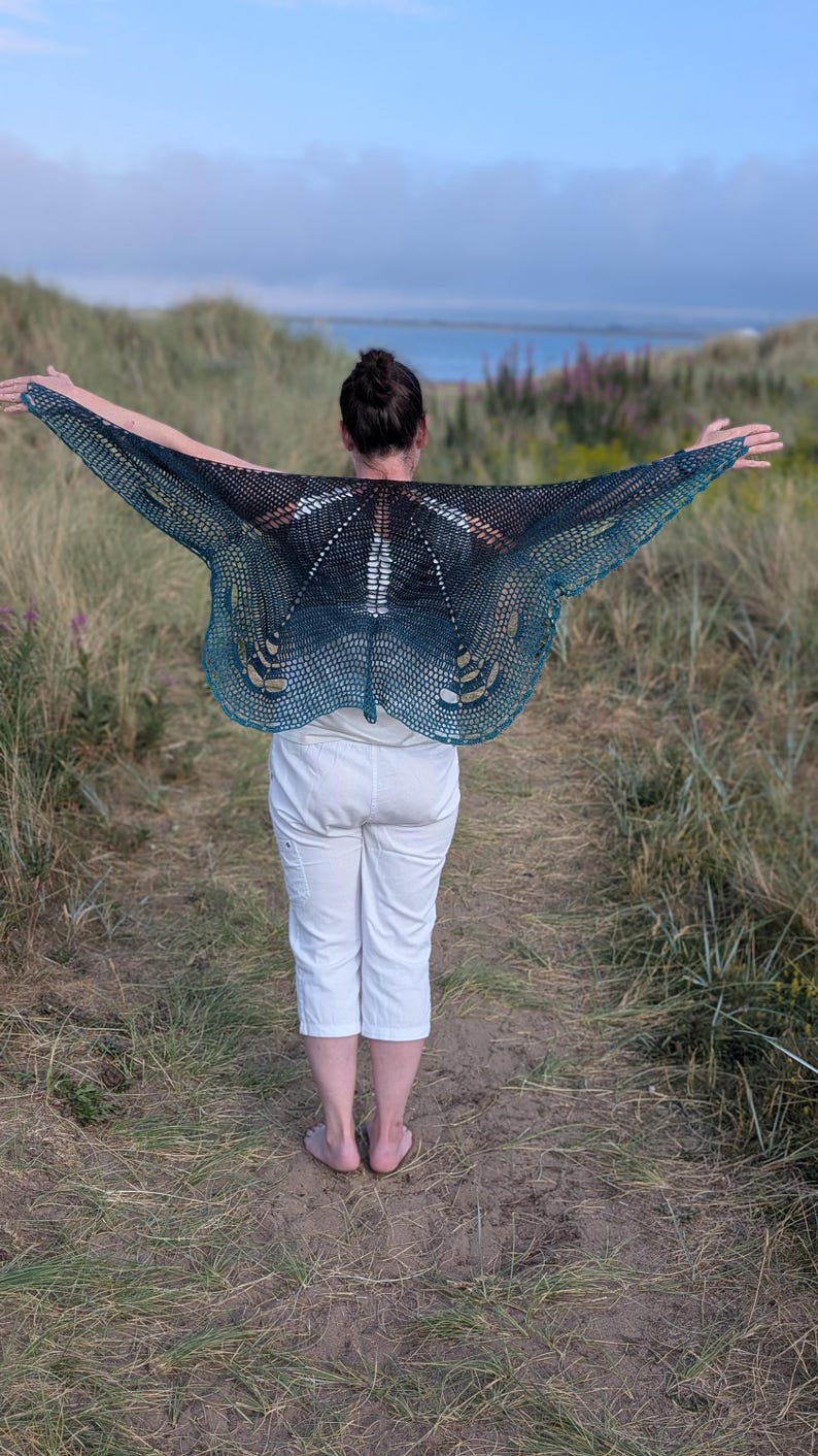 Puede incluir: Una persona lleva un chal hecho a mano, de tejido abierto, con un dise&ntilde;o de mariposa. El chal hace una transici&oacute;n de negro a verde azulado, con un patr&oacute;n delicado y calado. El fondo presenta un paisaje costero con dunas y el mar.