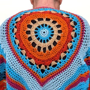 Op de afbeelding: Een kleurrijke, handgemaakte gehaakte trui met een groot cirkelvormig ontwerp op de rug. De trui heeft een levendig palet van blauw, oranje, roze en beige. Het open weefselontwerp is zichtbaar.