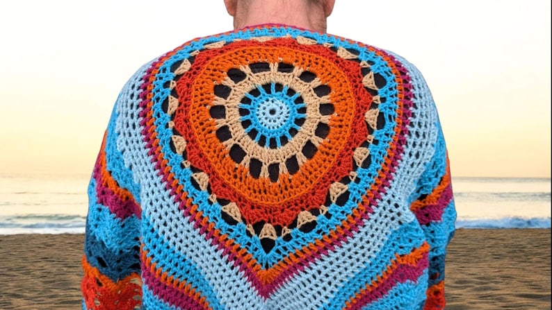 Op de afbeelding: Een kleurrijke, handgemaakte gehaakte trui met een groot cirkelvormig ontwerp op de rug. De trui heeft een levendig palet van blauw, oranje, roze en beige. Het open weefselontwerp is zichtbaar.