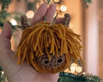Può includere: Un ornamento all'uncinetto a forma di testa di mucca delle Highlands. L'ornamento è marrone e giallo con una consistenza soffice e simile a un filo. L'ornamento è tenuto nella mano di una persona.