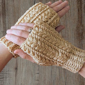 May include: A pair of beige crochet fingerless gloves. The gloves have a textured, woven pattern and are designed to cover the hand up to the knuckles. The gloves are shown on a wooden surface.