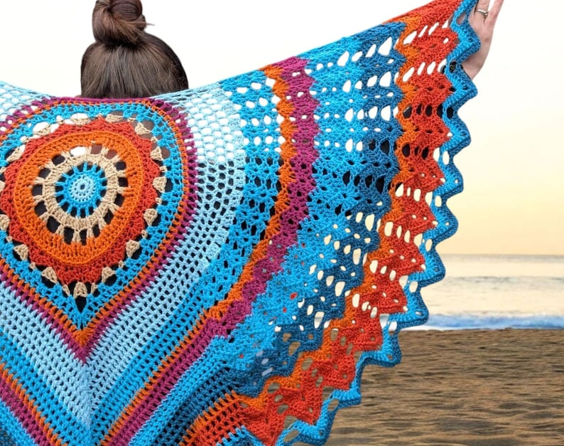 Op de afbeelding: Een kleurrijke, handgemaakte gehaakte sjaal met een levendig ontwerp. De sjaal heeft een centraal cirkelvormig patroon in oranje, beige en blauw, omgeven door lagen blauw, roze en oranje. De randen hebben een geschulpte vormgeving.