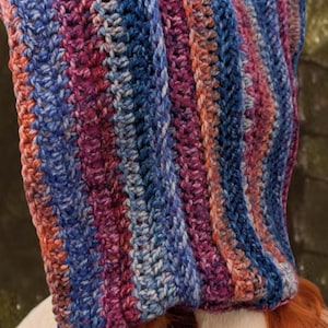 May include: A crocheted hooded scarf with a striped pattern in shades of blue, pink, and white. The scarf is made with a soft yarn and has a long fringe.