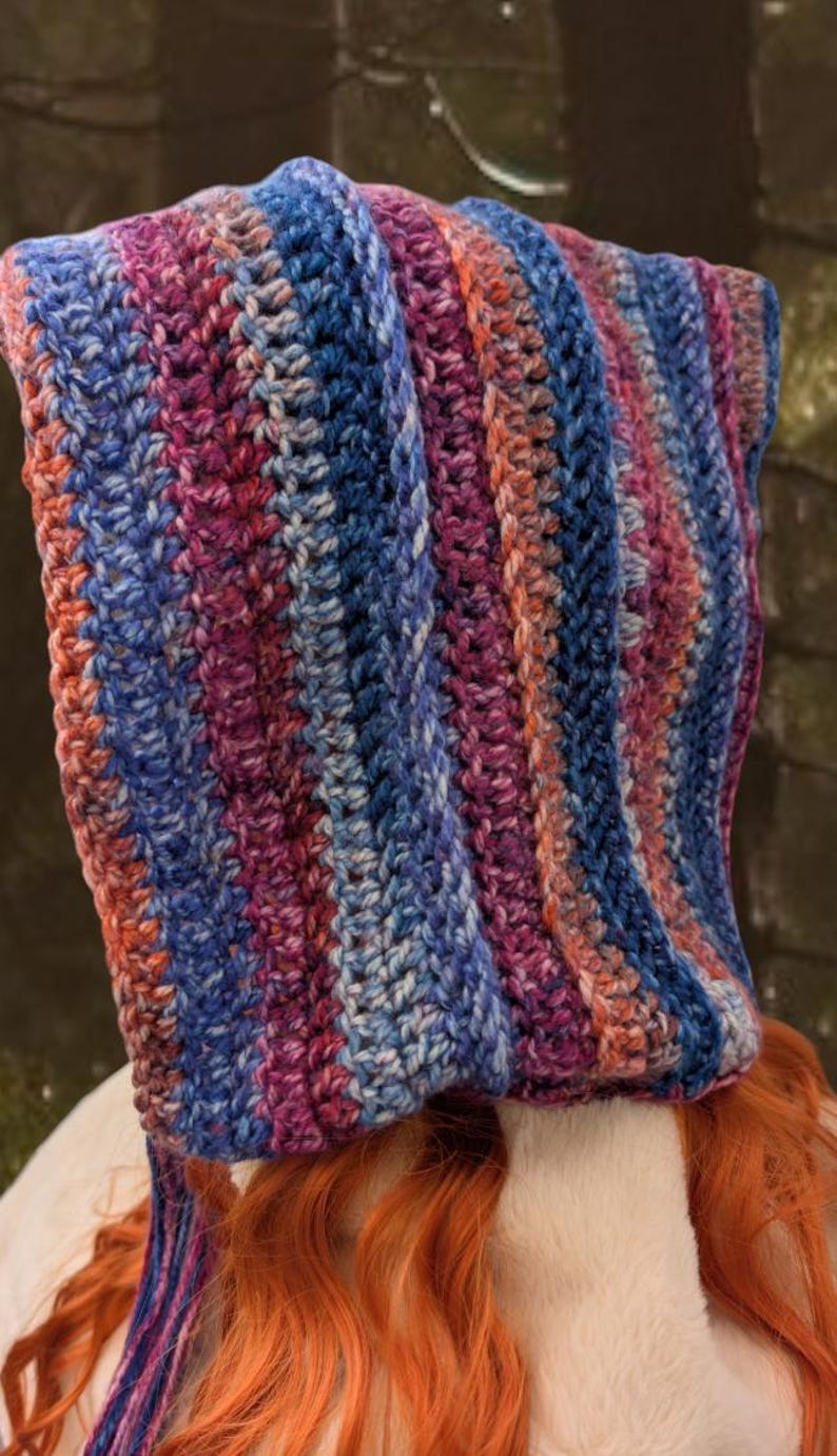 May include: A crocheted hooded scarf with a striped pattern in shades of blue, pink, and white. The scarf is made with a soft yarn and has a long fringe.