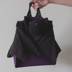 May include: A handmade crochet bat-shaped bag in black and purple. The bag features a bat head and wings, with a purple interior. The bag is held by a person's hand, showcasing the handle and overall design. The bag is made of a soft, textured yarn.