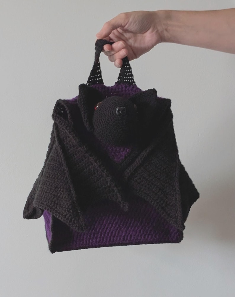 May include: A handmade crochet bat-shaped bag in black and purple. The bag features a bat head and wings, with a purple interior. The bag is held by a person's hand, showcasing the handle and overall design. The bag is made of a soft, textured yarn.