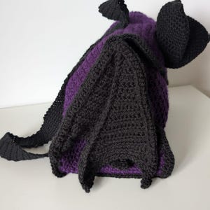 May include: A handmade crochet bat-shaped bag in black and purple. The bag features large, pointed ears and wings, with a strap for carrying. The body of the bag is purple, and the wings and ears are black.