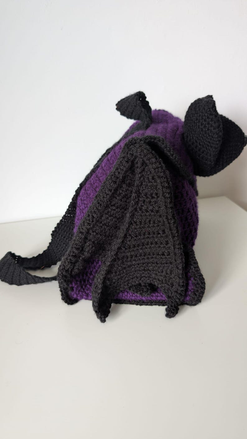 May include: A handmade crochet bat-shaped bag in black and purple. The bag features large, pointed ears and wings, with a strap for carrying. The body of the bag is purple, and the wings and ears are black.