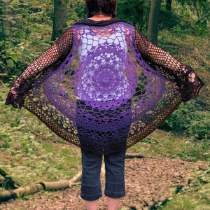 Puede incluir: Un chal de ganchillo hecho a mano en tonos morados y negros. El chal presenta un diseño de mandala circular en el centro, con un efecto degradado de morado claro a negro. El tejido abierto crea un aspecto delicado y aireado. El chal se lleva sobre una camiseta blanca y vaqueros azules.