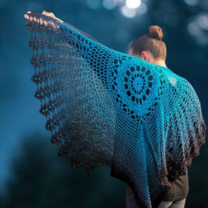 May include: A handmade crochet shawl in shades of blue and black. The shawl features a circular design on the back and a scalloped edge. The gradient color effect transitions from a bright blue to a dark black.