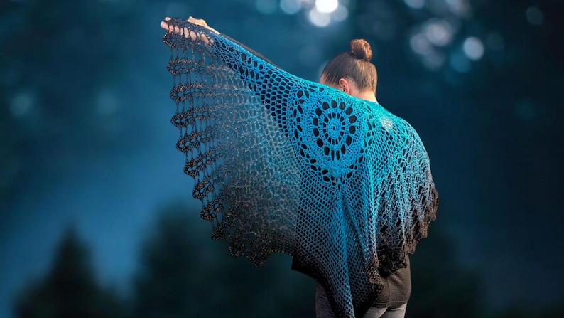 May include: A handmade crochet shawl in shades of blue and black. The shawl features a circular design on the back and a scalloped edge. The gradient color effect transitions from a bright blue to a dark black.