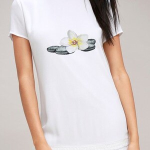 May include: White short-sleeve dress with a round neckline and a tiered, fringed hem. The dress features a watercolor print of a white and yellow flower resting on gray stones. A casual, summery style.