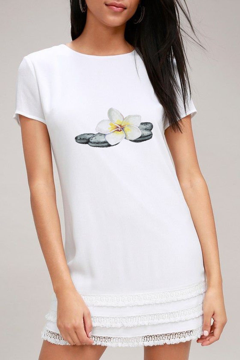 May include: White short-sleeve dress with a round neckline and a tiered, fringed hem. The dress features a watercolor print of a white and yellow flower resting on gray stones. A casual, summery style.