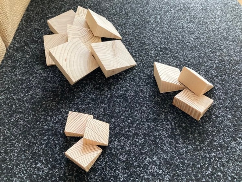 May include: A collection of small, unfinished wooden blocks. The blocks are square with one corner cut off, revealing the wood grain. They are scattered on a dark gray textured surface.