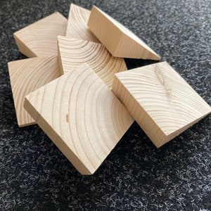 May include: A collection of unfinished, light-colored wooden blocks. Each block is square with a beveled edge, revealing the wood grain. The blocks are arranged in a slightly overlapping pattern, suggesting a craft or construction theme.