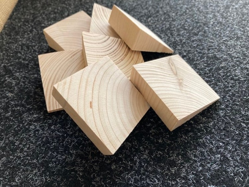 May include: A collection of unfinished, light-colored wooden blocks. Each block is square with a beveled edge, revealing the wood grain. The blocks are arranged in a slightly overlapping pattern, suggesting a craft or construction theme.