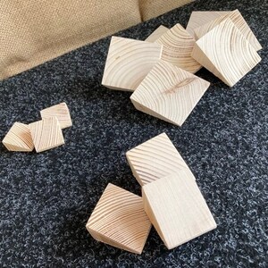 May include: A collection of unfinished, light-colored wooden blocks, each with visible wood grain patterns. The blocks are square with beveled edges, scattered on a dark gray textured surface. The blocks vary in size and are likely for a craft or building project.
