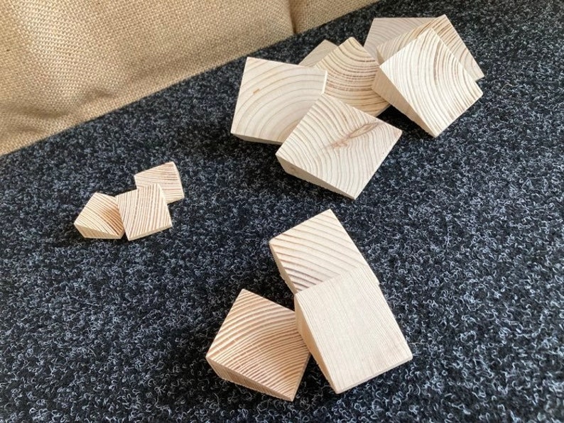 May include: A collection of unfinished, light-colored wooden blocks, each with visible wood grain patterns. The blocks are square with beveled edges, scattered on a dark gray textured surface. The blocks vary in size and are likely for a craft or building project.