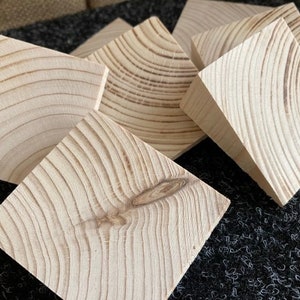 May include: A collection of six unfinished, square wooden blocks. The blocks are light-colored with visible wood grain patterns and knots. The blocks are arranged in a slightly overlapping manner, showcasing their natural texture and shape.
