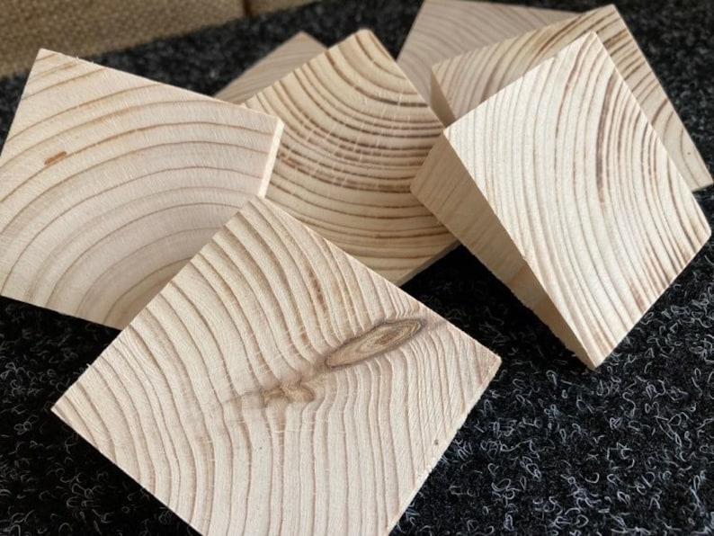 May include: A collection of six unfinished, square wooden blocks. The blocks are light-colored with visible wood grain patterns and knots. The blocks are arranged in a slightly overlapping manner, showcasing their natural texture and shape.
