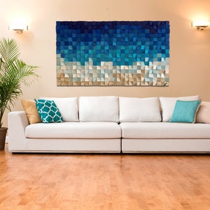 Op de afbeelding: Een witte bank met turquoise en beige kussens staat voor een muur met een blauw en beige geometrisch wandkunstwerk.
