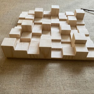 May include: A wooden wall panel made of various sized blocks, creating a textured surface. The panel is light brown and has a natural wood grain.