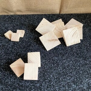 May include: A collection of unfinished, square wooden blocks with visible wood grain. The blocks are arranged on a dark, textured surface, with a neutral-colored backdrop. The blocks vary in size and are likely for crafts or building.