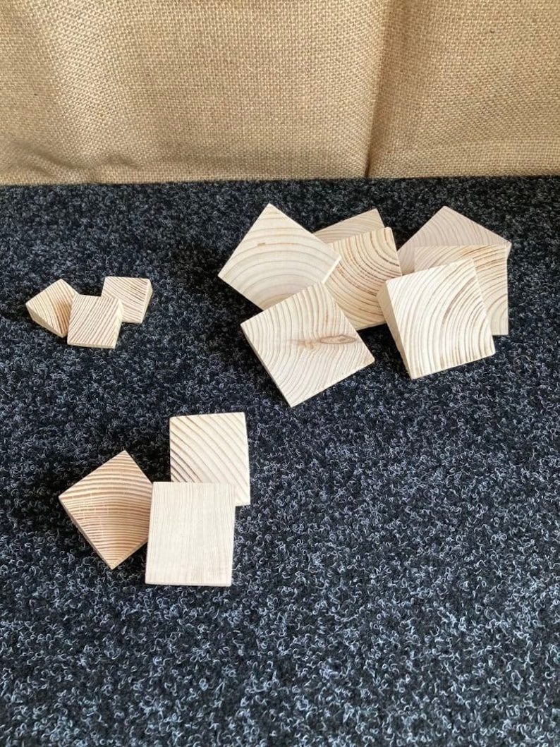 May include: A collection of unfinished, square wooden blocks with visible wood grain. The blocks are arranged on a dark, textured surface, with a neutral-colored backdrop. The blocks vary in size and are likely for crafts or building.
