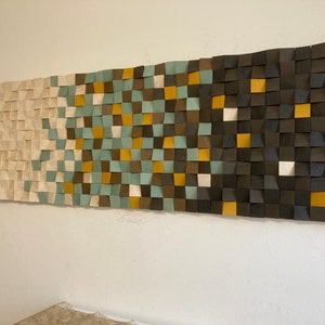 Pu&ograve; includere: Un'opera d'arte da parete realizzata con quadrati di legno in varie tonalit&agrave; di marrone, beige, verde acqua e oro. I quadrati sono disposti a forma di onda, creando un design moderno e astratto.