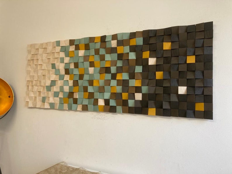 Pu&ograve; includere: Un'opera d'arte da parete realizzata con quadrati di legno in varie tonalit&agrave; di marrone, beige, verde acqua e oro. I quadrati sono disposti a forma di onda, creando un design moderno e astratto.