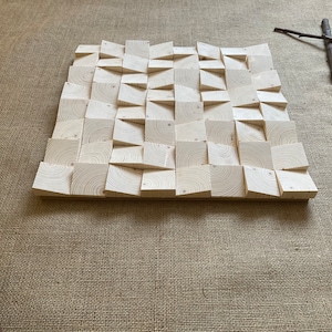 May include: A square wall panel made of light wood with a geometric pattern. The panel is made up of many small squares and triangles, all of which are slightly raised. The panel is a light wood color and has a natural finish.