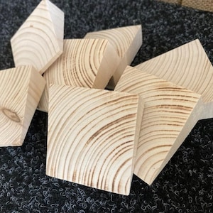 Op de afbeelding: Een verzameling onafgewerkte houten blokken in verschillende geometrische vormen. De blokken zijn gemaakt van lichtgekleurd hout, met zichtbare nerfpatronen en knopen. Sommige blokken zijn vierkant, andere hebben schuine zijden. De blokken liggen verspreid op een donkere ondergrond.