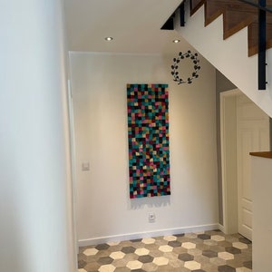 May include: A colorful geometric wall hanging made of wood blocks in shades of pink, teal, yellow, and brown. The wall hanging is mounted on a white wall in a hallway with a hexagonal tile floor.