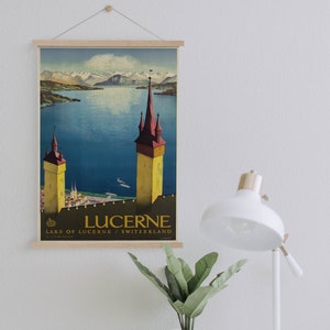 May include: A vintage travel poster for Lucerne, Switzerland, featuring a lake scene with two tall towers and snow-capped mountains in the background. The poster is framed with a wooden hanger and displays the word "LUCERNE".