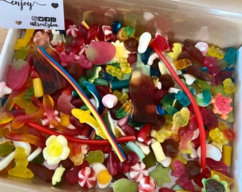 May include: A box overflowing with a colorful assortment of gummy candies. The mix includes gummy worms, cola bottle shapes, fruit slices, and other assorted shapes and colors. A small card in the corner reads "enjoy" with social media handles.