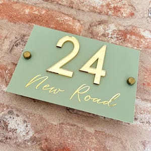 Op de afbeelding: Een huisnummerbord in saliegroen met gouden cijfers "24" en de tekst "New Road" in gouden schrift.