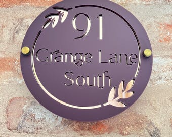 May include: A round, plum-colored house number sign with the number "91" and the text "Grange Lane South" cut out. The sign has gold-colored accents and leaf designs. It is mounted on a brick wall.