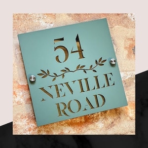 Puede incluir: Un letrero cuadrado de número de casa azul verdoso con el número 54 y el nombre de la calle Neville Road recortado en el letrero. El letrero tiene un diseño decorativo de vid.