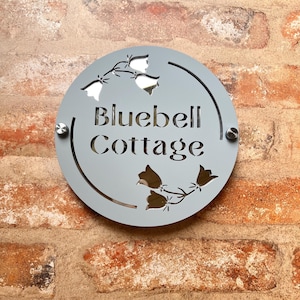 Op de afbeelding: Een ronde, lichtgrijze metalen plaat met de woorden "Bluebell Cottage" uitgesneden. Het bord heeft bloemmotieven en is met twee zilveren schroeven aan een bakstenen muur bevestigd.
