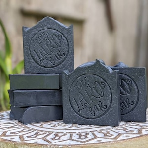 May include: A stack of five charcoal soap bars with the text "The Laro Bar" embossed on each bar. The soap bars are arranged on a wooden surface with a decorative pattern.