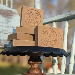May include: A stack of four brown soap bars with the text "The Laro Bar" embossed on each bar. The soap bars are displayed on a black cake stand with a wooden base.