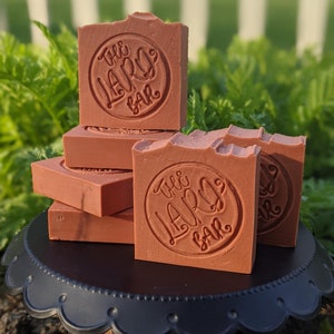 May include: A stack of five red-brown soap bars with the words "The Lard Bar" embossed on the top bar. The soap bars are on a black cake stand with a scalloped edge.