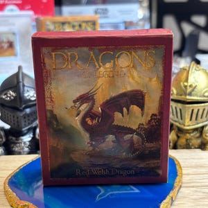 May include: A red box featuring the text "DRAGONS OF LEGEND" and a painting of a red dragon. The text "Red Welsh Dragon" is below the image. The box is on a blue and gold decorative coaster.