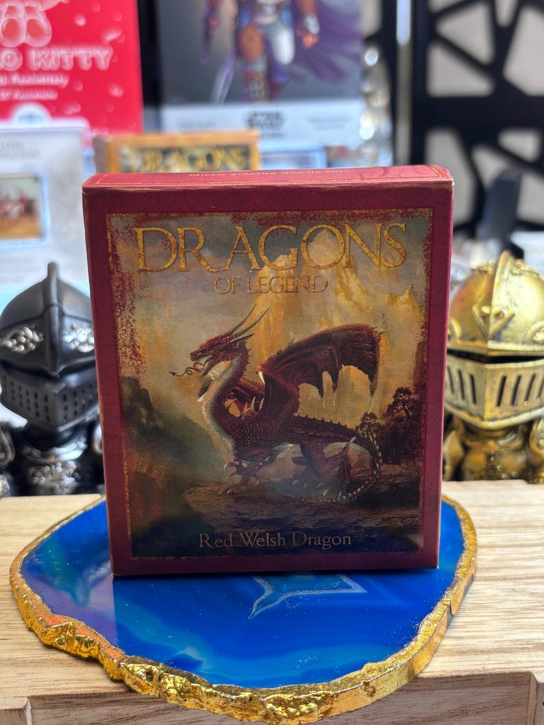 May include: A red box featuring the text "DRAGONS OF LEGEND" and a painting of a red dragon. The text "Red Welsh Dragon" is below the image. The box is on a blue and gold decorative coaster.
