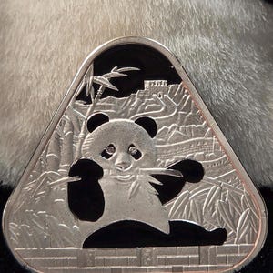 May include: A triangular silver-toned coin featuring a relief of a panda eating bamboo. The background depicts the Great Wall of China and bamboo stalks against a black enamel backdrop. The coin has a metallic border and a textured base design.