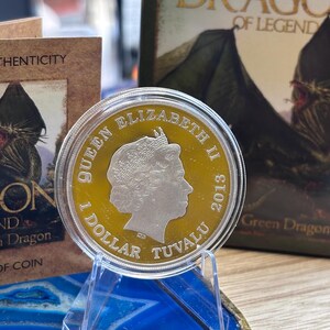 May include: A silver and gold coin featuring Queen Elizabeth II, dated 2013, with the text "1 DOLLAR TUVALU". The coin is displayed on a clear acrylic stand, with a blue and gold agate slice in the foreground and a book titled "DRAGO OF LEGEND" in the background.