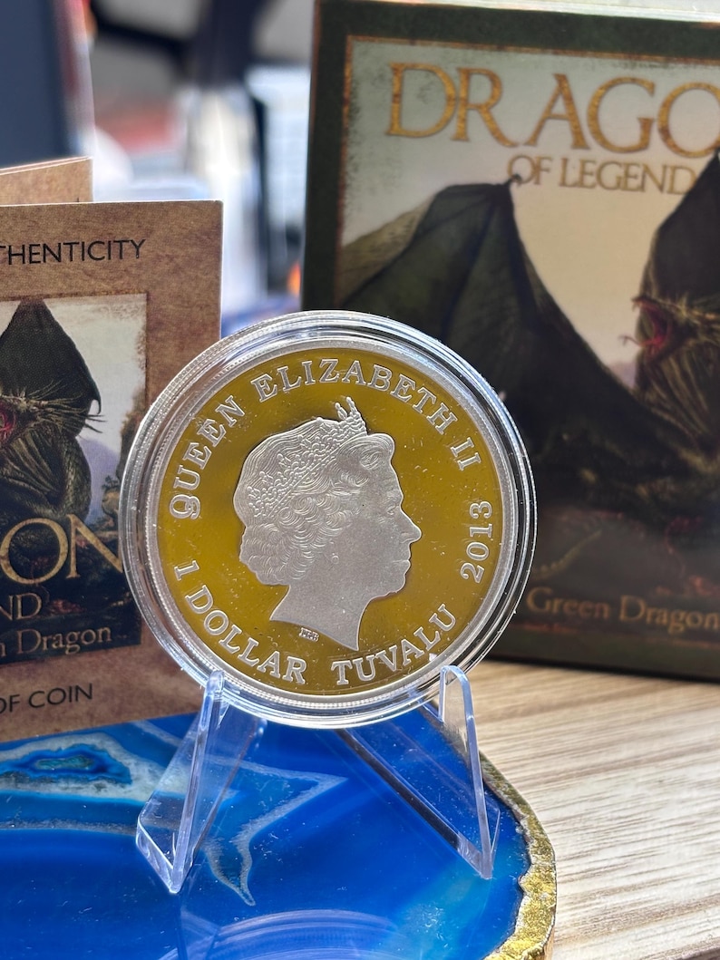 May include: A silver and gold coin featuring Queen Elizabeth II, dated 2013, with the text "1 DOLLAR TUVALU". The coin is displayed on a clear acrylic stand, with a blue and gold agate slice in the foreground and a book titled "DRAGO OF LEGEND" in the background.